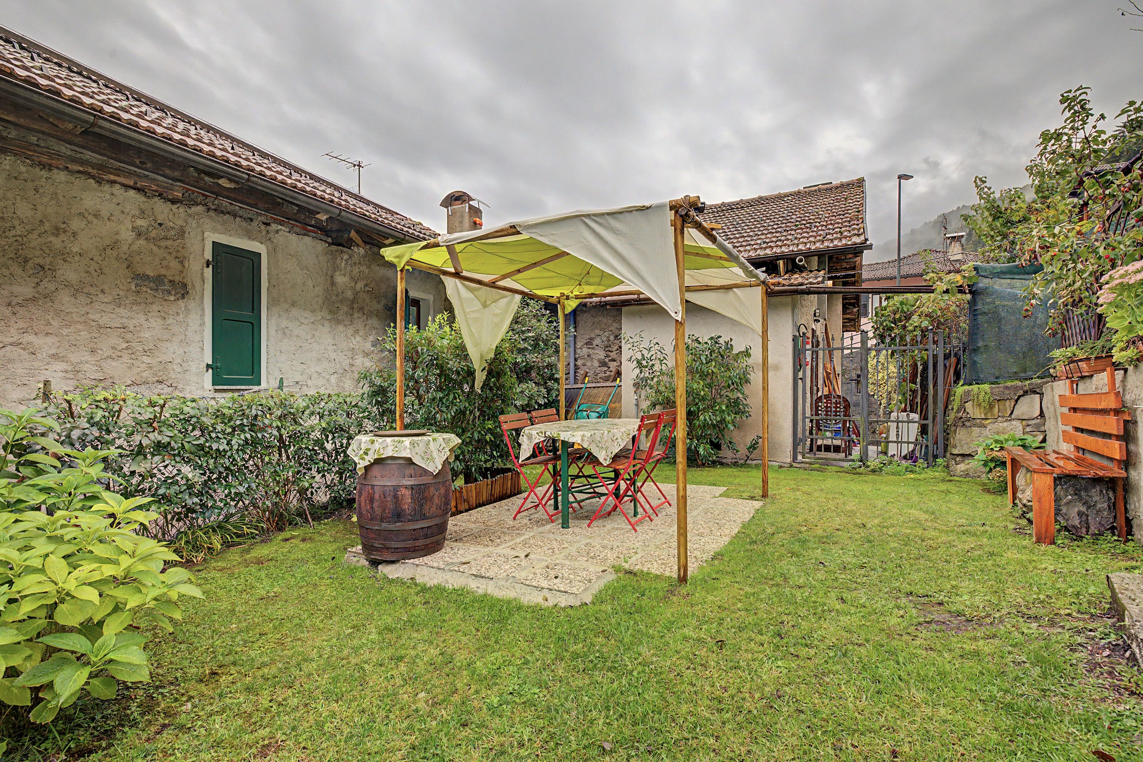 Un área de estar al aire libre con estilo rústico, con una mesa, sillas bajo un toldo sombreado, un barril de madera y un muro de piedra.