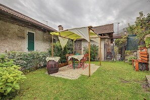A rustic outdoor seating area with a table and chairs under a shaded canopy, a wooden barrel, and a stone wall.