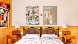 Habitación de hotel con cabecera de madera, cama grande con cubrecama estampado, dos mesitas de noche con lámparas, estantería con libros y dos carteles enmarcados en la pared.