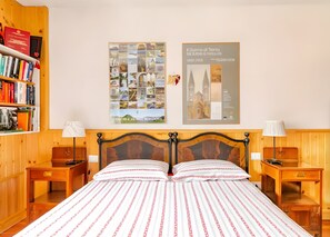 Une chambre d’hôtel avec une tête de lit en bois, un grand lit recouvert d’une couvre-lit à motifs, deux tables de chevet avec des lampes, une bibliothèque remplie de livres et deux affiches encadrées accrochées au mur.