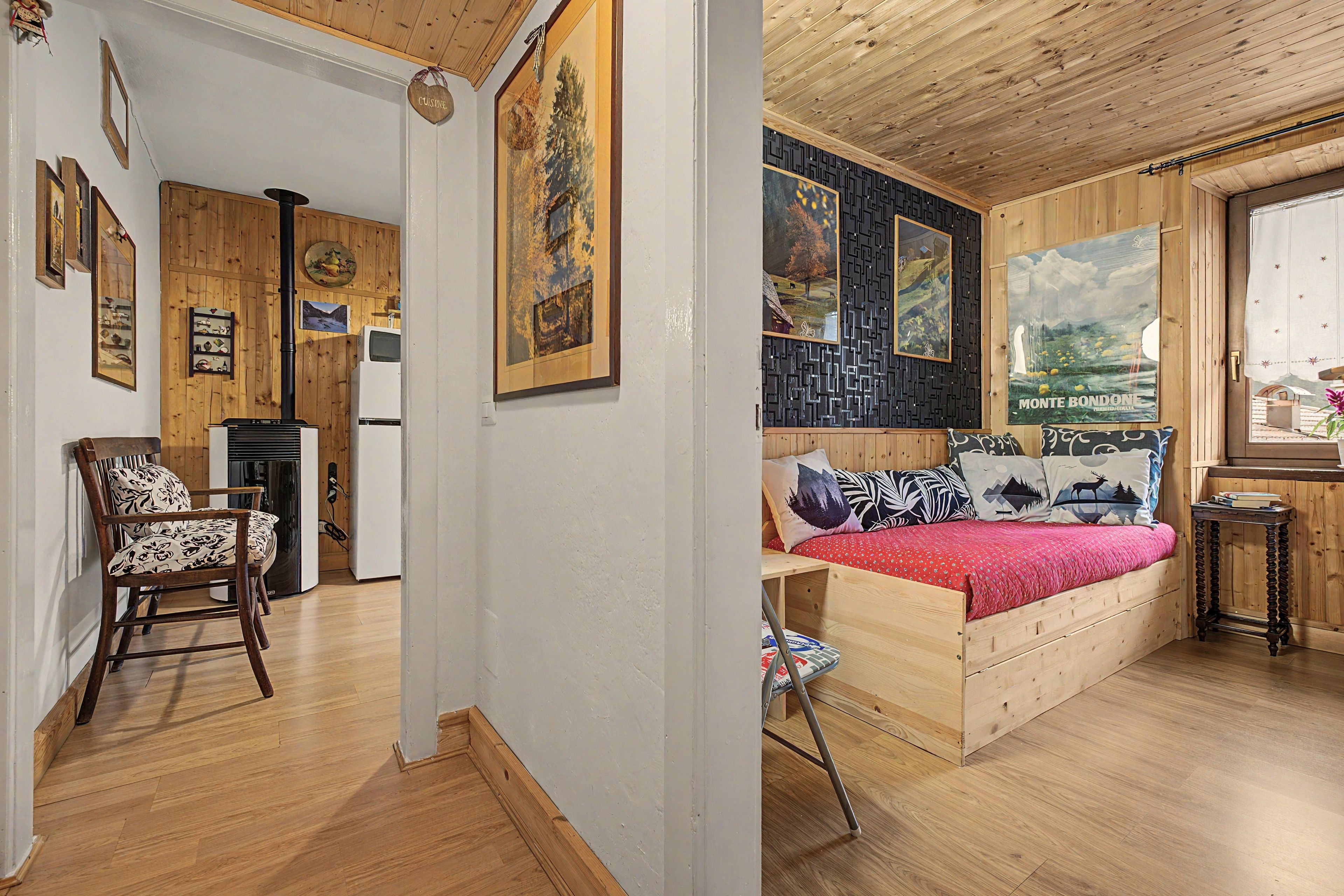 Ein Zimmer mit einem Bett, einem Sessel, einem kleinen Tisch und einem Fenster. Die Wände und Decke sind aus Holz.