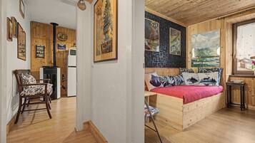 Ein Zimmer mit einem Bett, einem Sessel, einem kleinen Tisch und einem Fenster. Die Wände und Decke sind aus Holz.