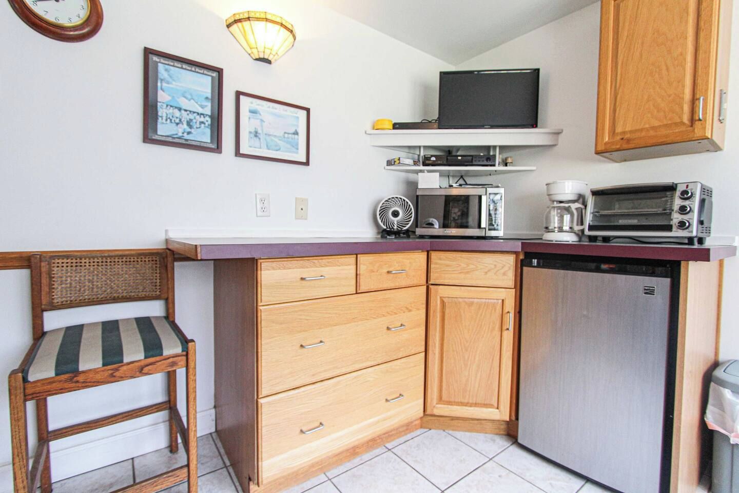 Une cuisine compacte équipée d’un four à micro-ondes, d’un grille-pain et d’un petit réfrigérateur. On y trouve une chaise en bois avec un coussin à rayures et une étagère fixée au mur, au-dessus du plan de travail.