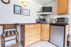 A compact kitchen with a microwave, toaster, and a small refrigerator. There is a wooden chair with a striped cushion and a wall-mounted shelf above the counter. - A budget-friendly, freshly renovated studio at Sunnyside, just steps from the lake. (Greenbush)