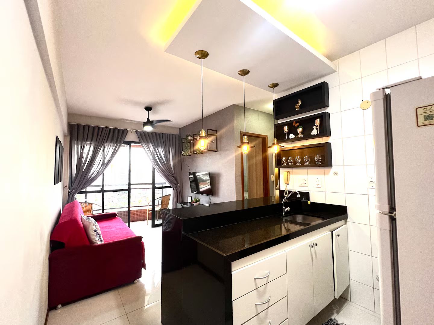 A modern kitchen with a black countertop, white cabinets, and a built-in oven. There is a red sofa in the adjacent living area, and a ceiling fan above.