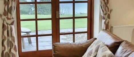 A room with a brown leather sofa, a view of a grassy area through a window with curtains, and a white radiator on the wall.