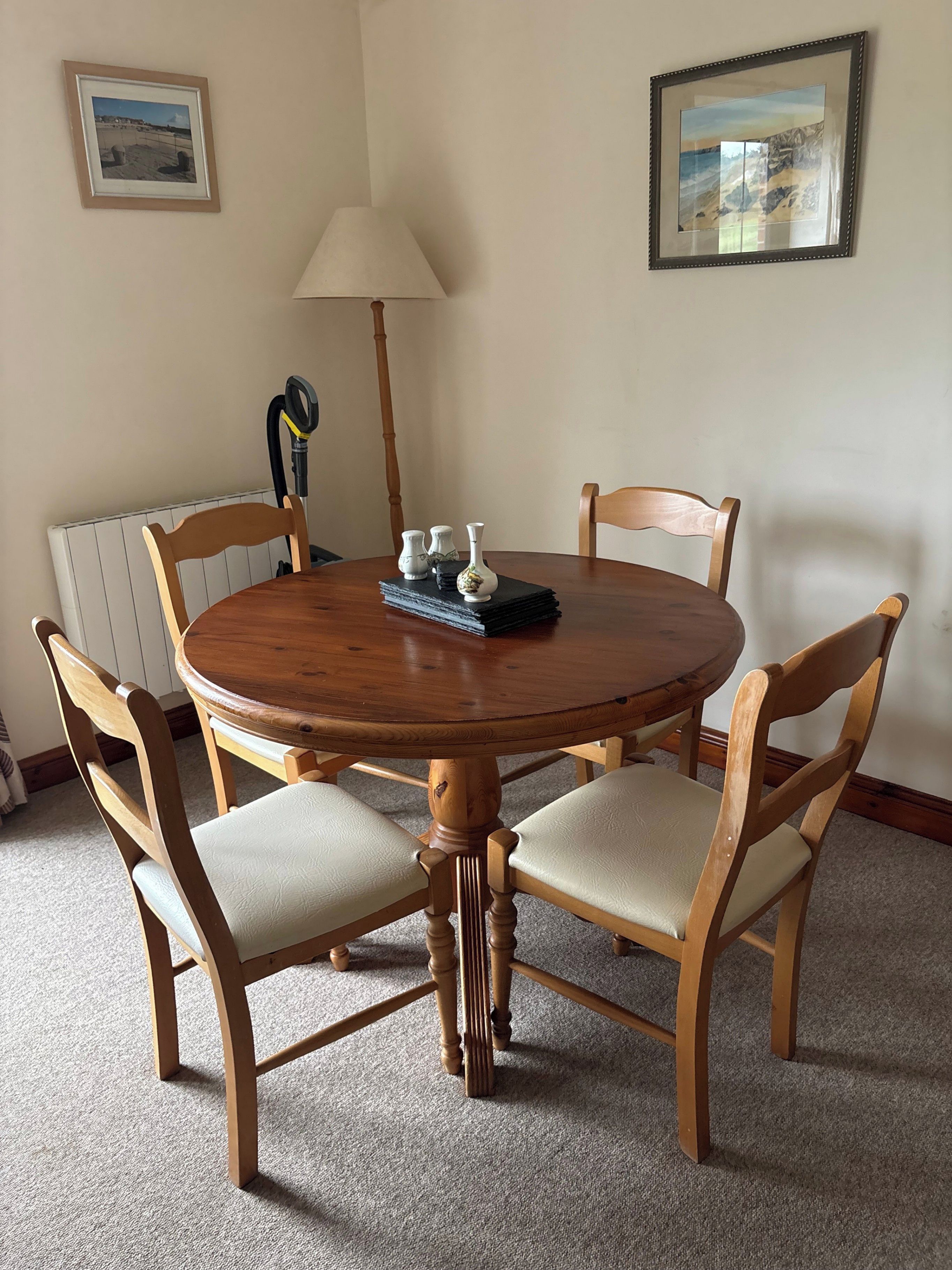 A round wooden dining table with four chairs, a lamp, and two framed pictures on the wall.