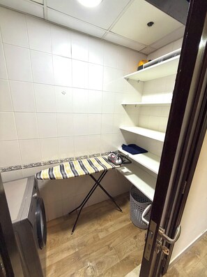 A laundry area with a washing machine, an ironing board, and shelves. - Homeland Female Hostel (Dubai)