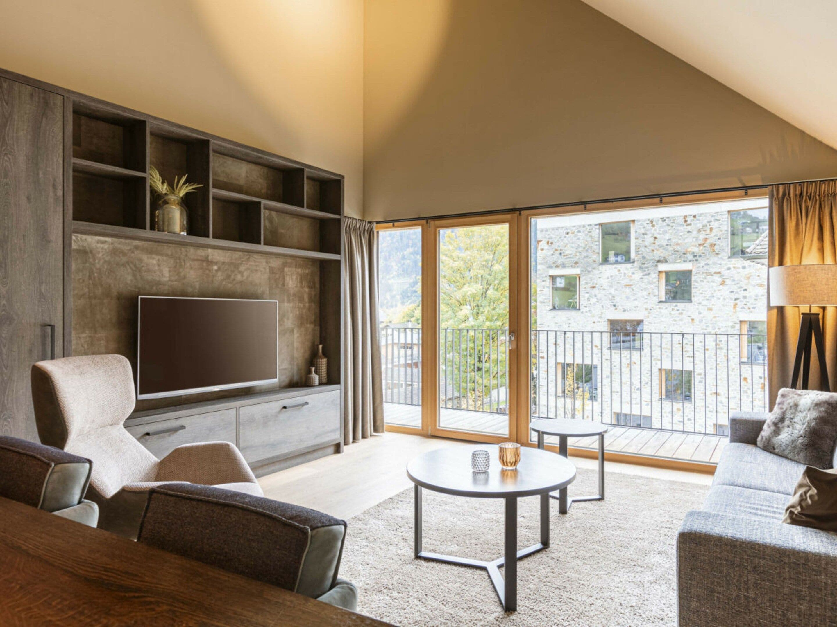 Una sala moderna con una televisión de pantalla plana, estanterías empotradas, un sofá, una mesa de centro y un balcón con vistas a un edificio de piedra.