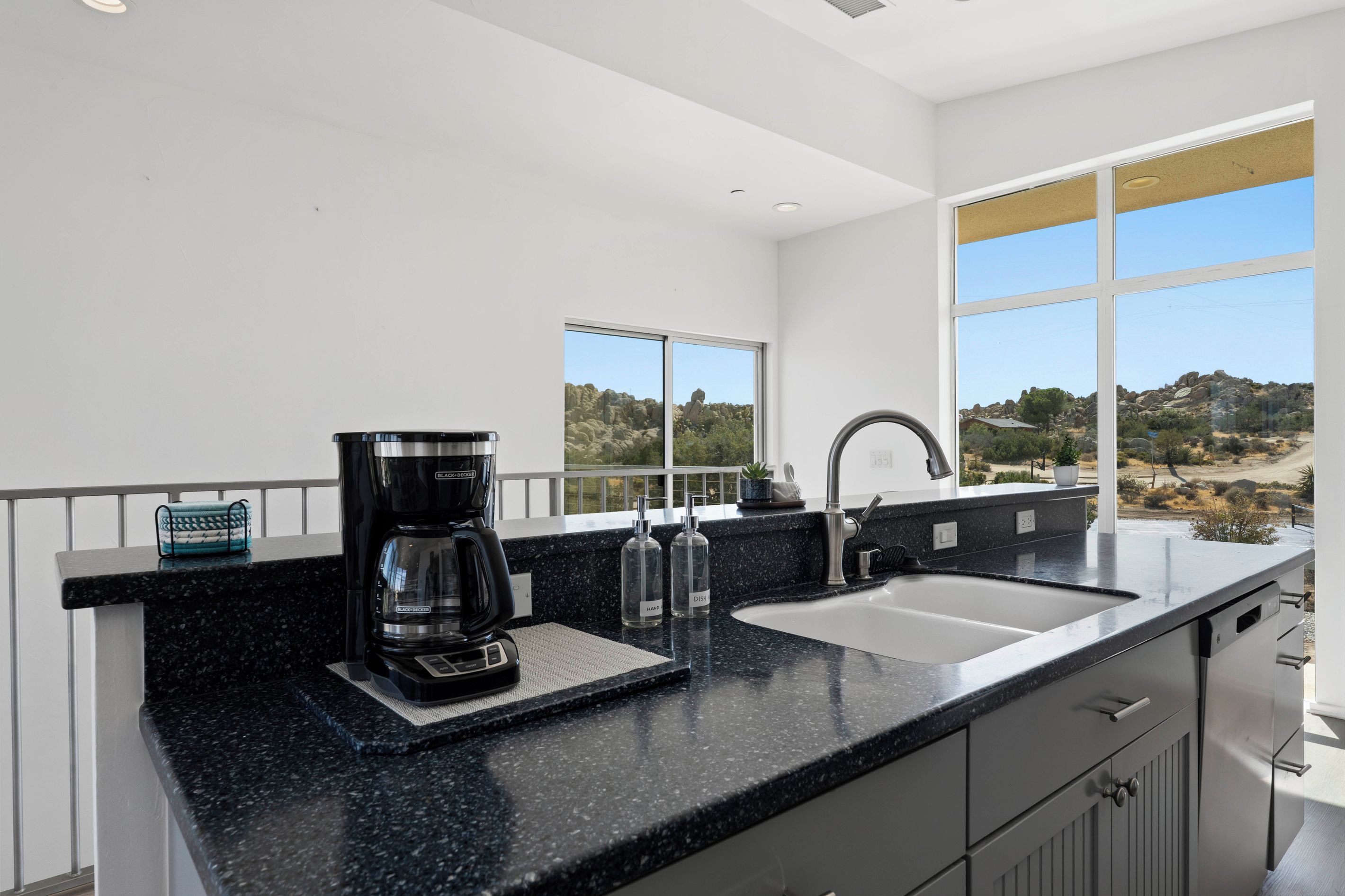 Cozinha moderna com bancada de granito preto, cafeteira, pia e vista para uma paisagem rochosa através de grandes janelas.