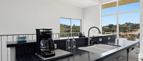 A modern kitchen with a black granite countertop, a coffee maker, a sink, and a view of a rocky landscape through large windows.