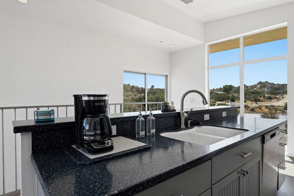 Eine moderne Küche mit schwarzer Granitabdeckung, einer Kaffeemaschine, einem Spülbecken und Blick auf eine felsige Landschaft durch große Fenster.