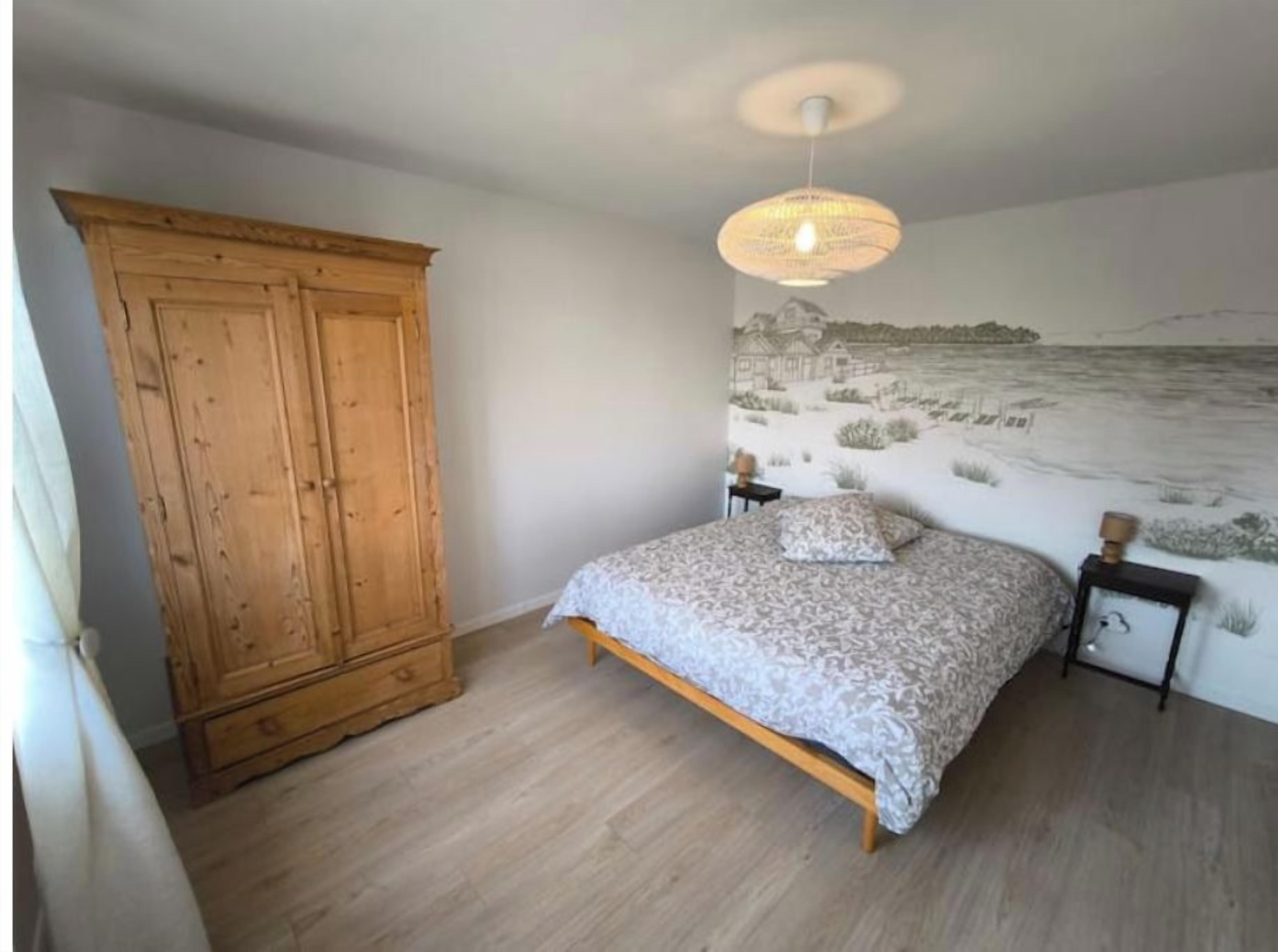 A bedroom with a bed, a wooden wardrobe, a wall mural, and a bedside table with a lamp.
