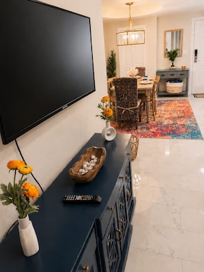 Ein modernes Wohnzimmer mit einem Flachbildfernseher, einem Holztische mit Dekorationen, einem Essbereich mit einem gemusterten Teppich und einer Chandelüster.