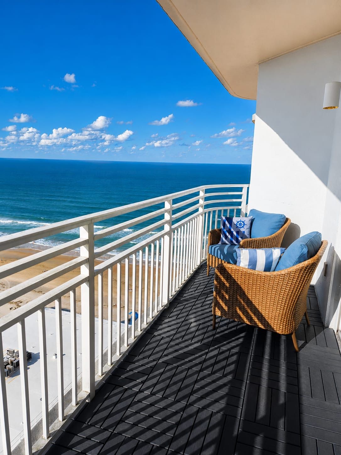 A balcony with a wicker chair and cushions, overlooking a beach and the ocean.