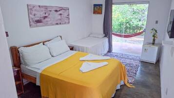 Quarto com cama, mesa de cabeceira, mesinha, rede e janela com vista para o verde.