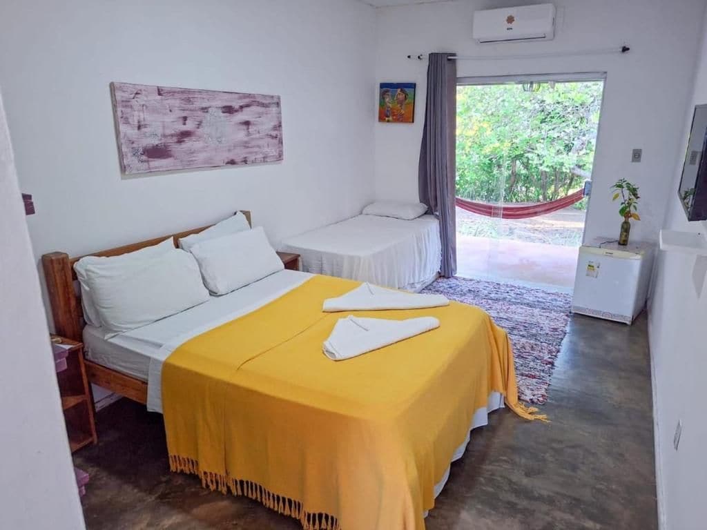 Un dormitorio con cama, mesita de noche, una pequeña mesa, una hamaca y una ventana con vistas a la vegetación.