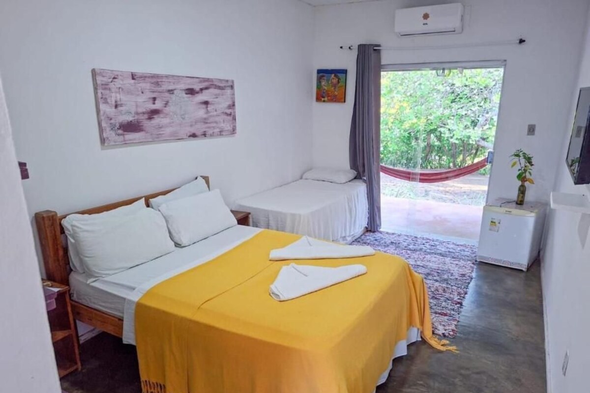 A bedroom with a bed, a bedside table, a small table, a hammock, and a window with a view of greenery.