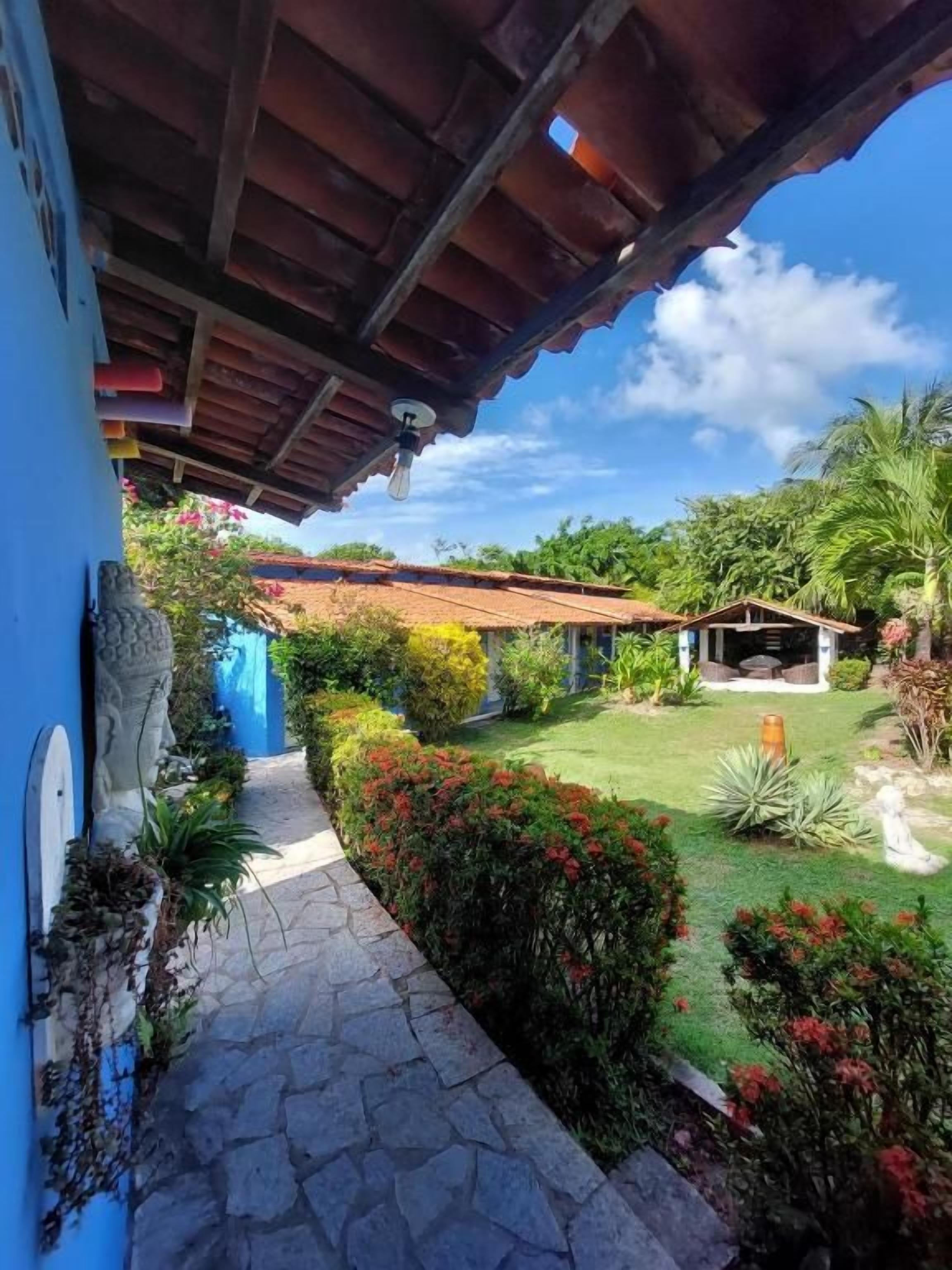 Uma trilha leva a uma casa com telhado de telhas vermelhas, rodeada por um jardim com diversas plantas e um céu claro.