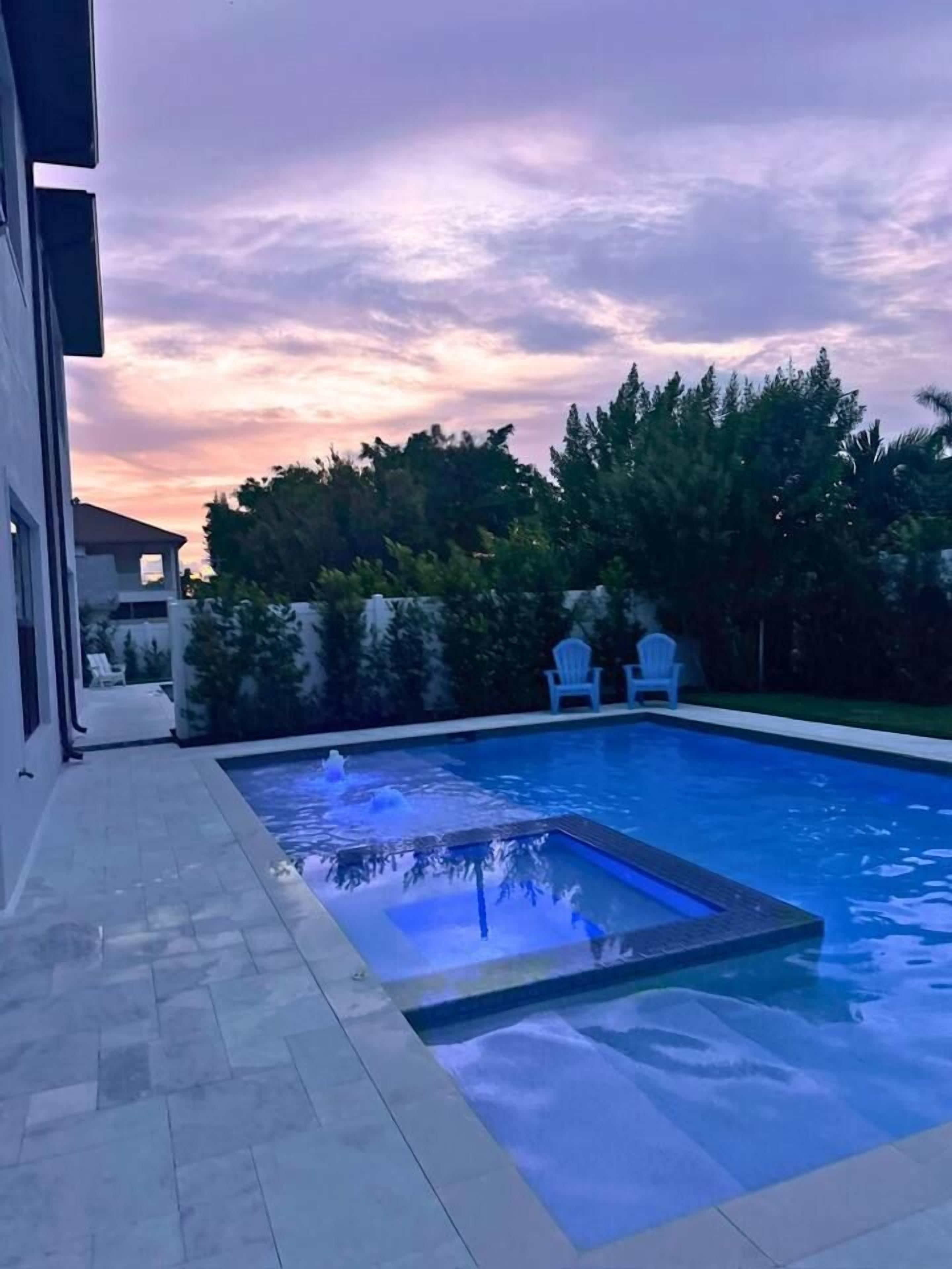 A swimming pool with a built-in hot tub, surrounded by a patio area with two chairs, and a backdrop of trees and a house.