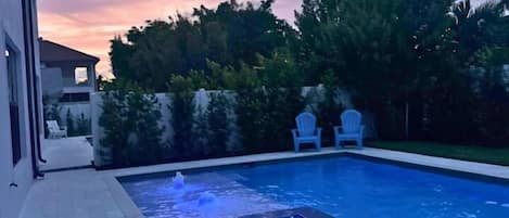 A swimming pool with a built-in hot tub, surrounded by a patio area with two chairs, and a backdrop of trees and a house.