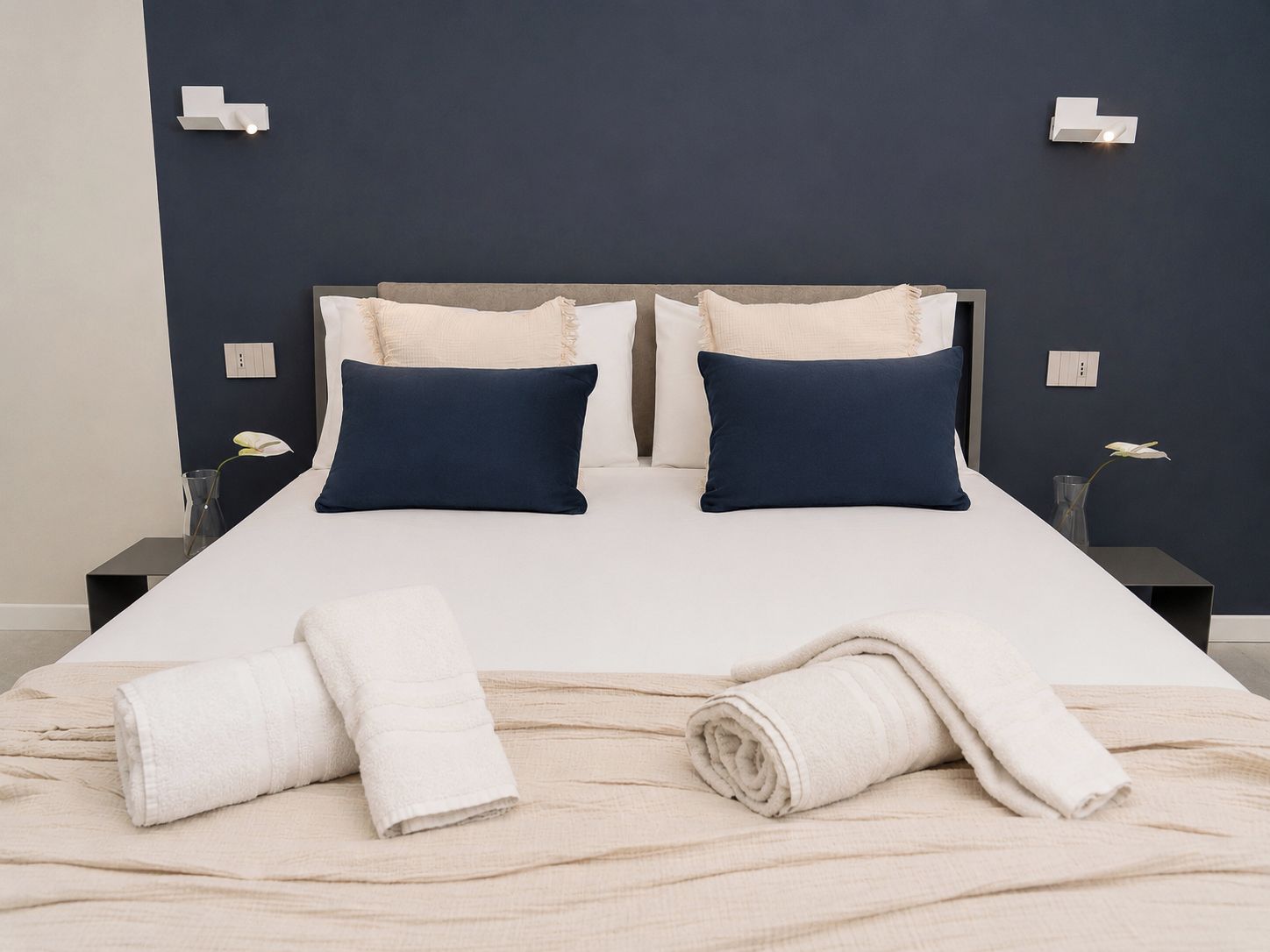 Een keurig opgemaakt bed met twee kussens, een nachtkastje met een telefoon, en een vaas met een bloem erop.