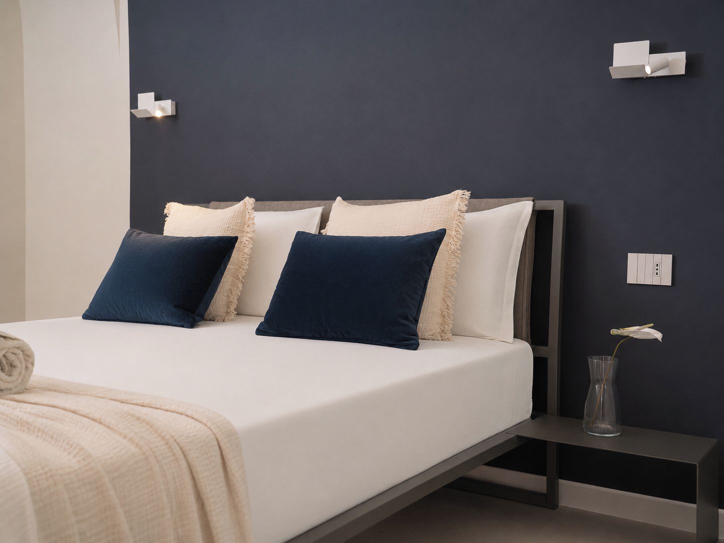 A modern bedroom with a bed, pillows, a bedside table, and a wall-mounted light fixture.