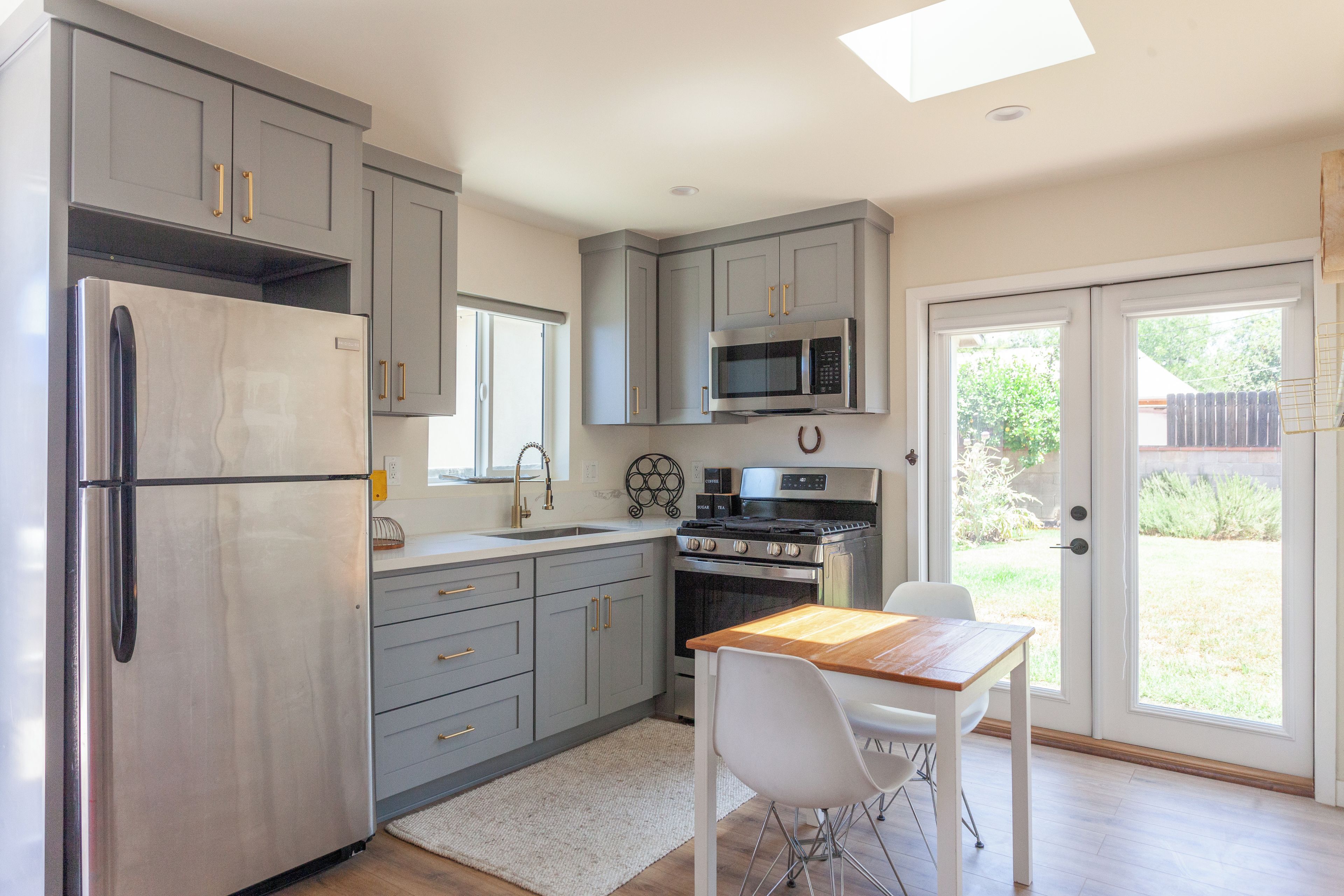 A modern kitchen with grey cabinets, a stainless steel refrigerator, a microwave, and an oven. There is a dining table with chairs and a window providing natural light.