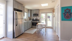 Una cocina moderna con electrodomésticos de acero inoxidable, una zona de comedor con mesa y silla blancos, y vistas a través de la puerta de cristal.