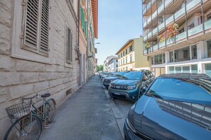 Eine enge Straße, gesäumt von parkenden Autos und einem Fahrrad. Häuser auf beiden Seiten haben Balkone mit Pflanzen.