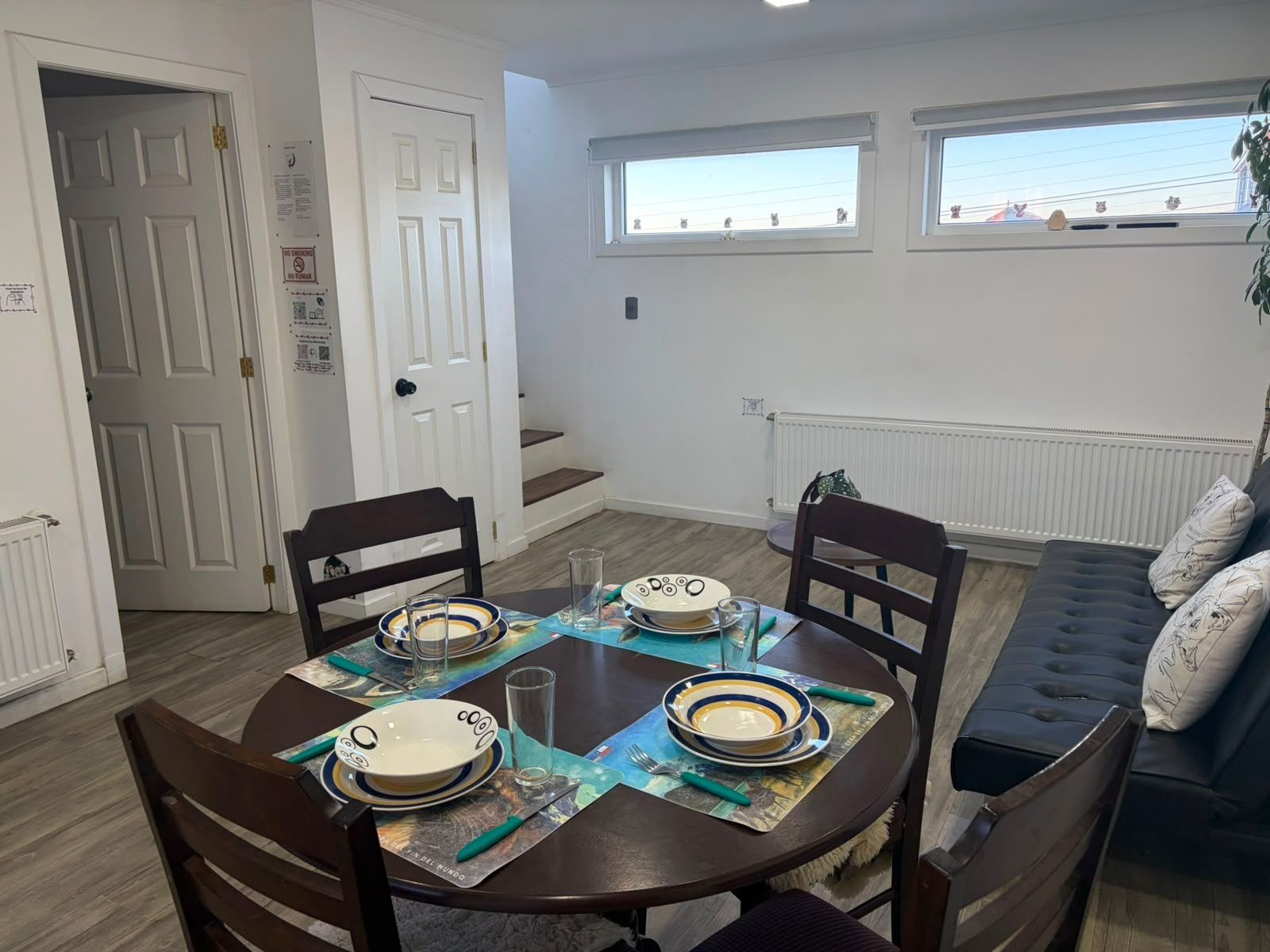 Uma sala de jantar com uma mesa redonda posta para quatro pessoas, cadeiras, um sofá e uma janela com vista.