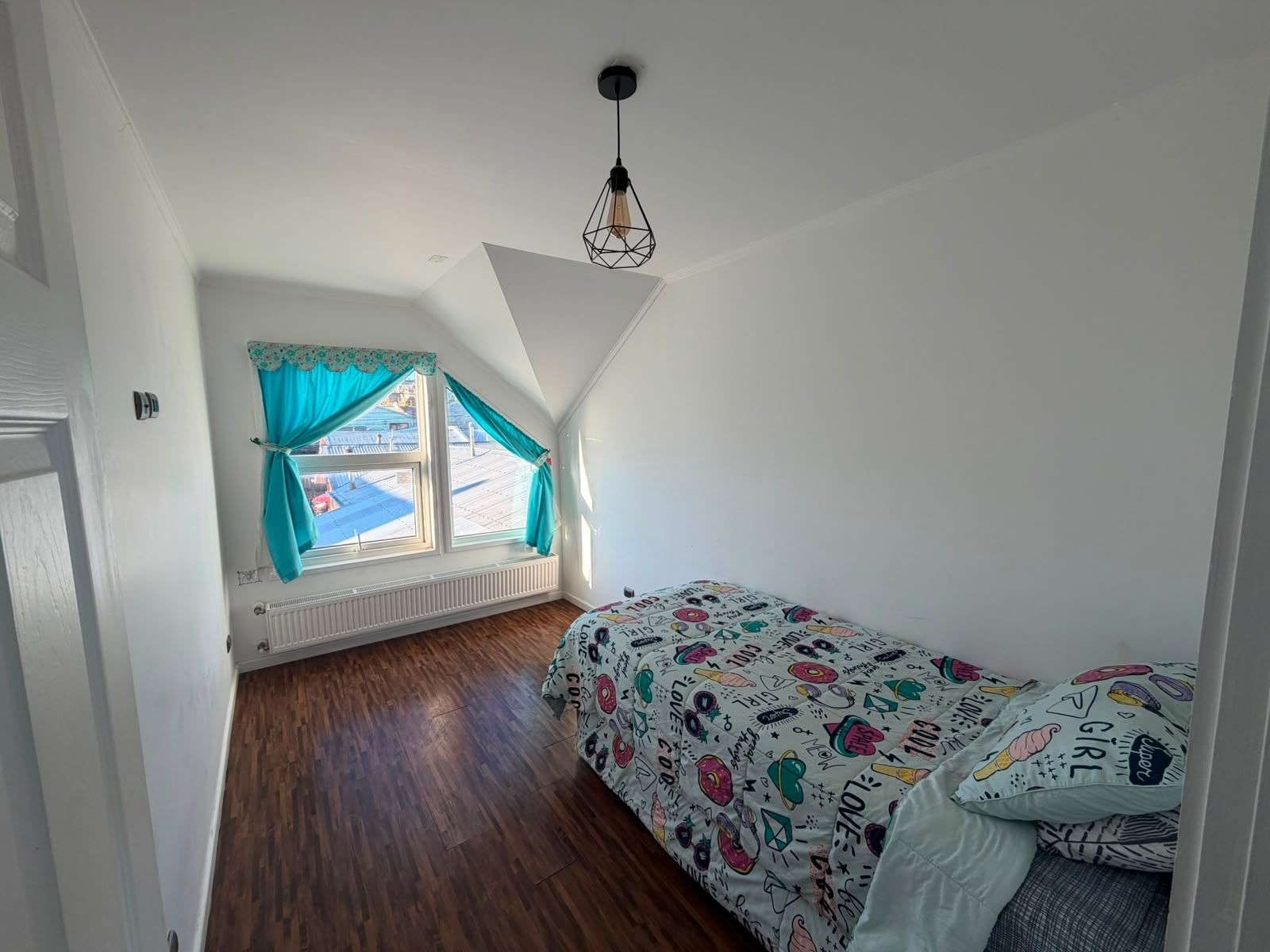 Una camera da letto con soffitto spiovente, una finestra con tende, un letto con biancheria a motivi e una lampada a sospensione.