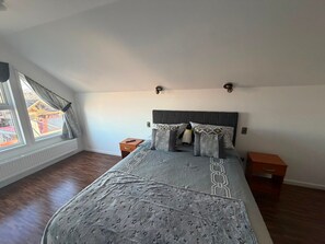 Un dormitorio con cama, dos mesitas de noche, una ventana con cortinas y vista a un edificio.