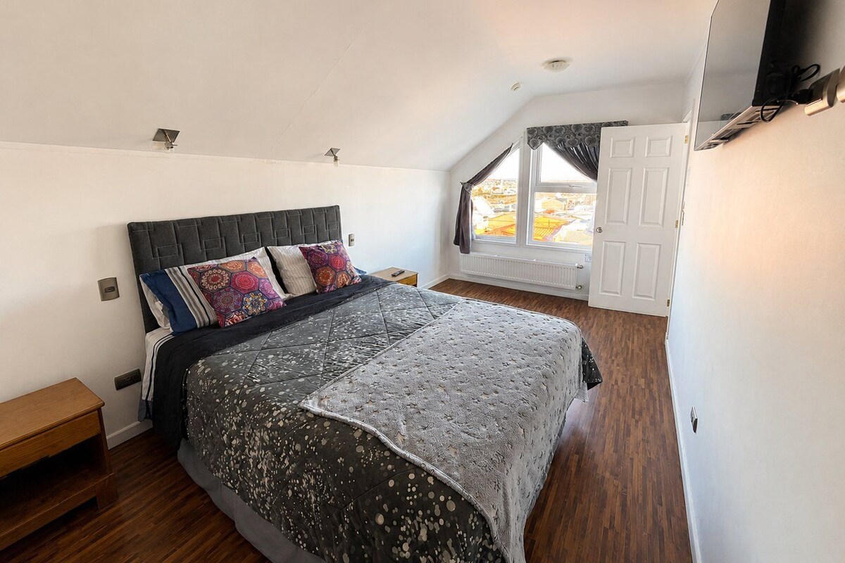 A bedroom with a large bed, a window with curtains, a wooden floor, and a white door.