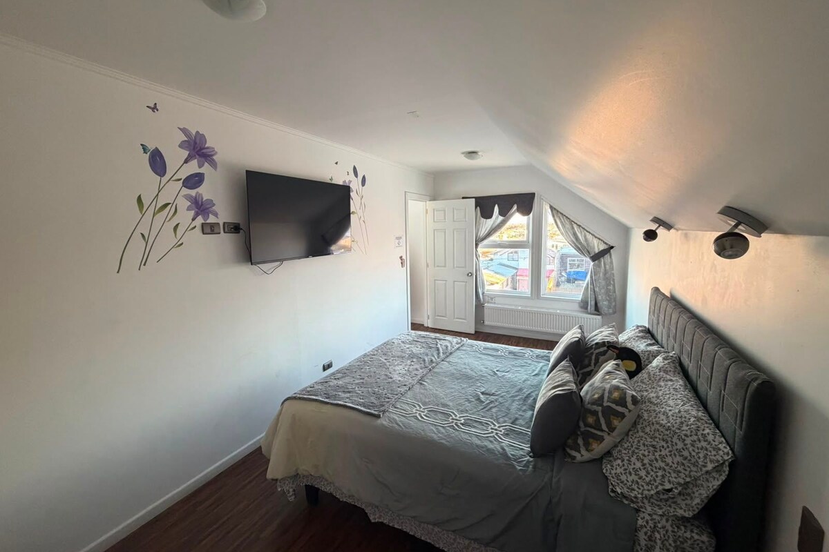 A bedroom with a bed, a flat-screen TV mounted on the wall, a window with curtains, and a ceiling light.