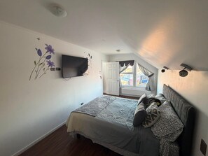 A bedroom with a bed, a flat-screen TV mounted on the wall, a window with curtains, and a ceiling light.