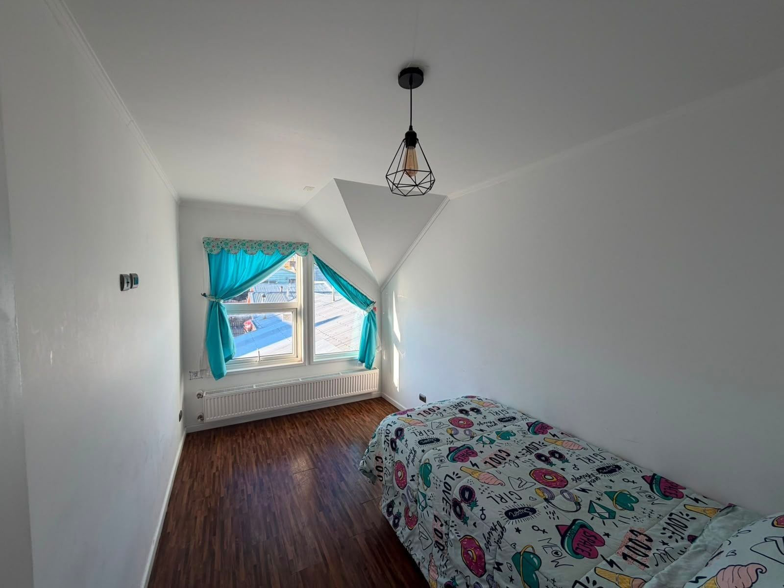 Un petit appartement d’une seule pièce, avec un plafond en pente, une fenêtre avec des rideaux, un radiateur et un lit recouvert d’une courtepointe aux couleurs vives.