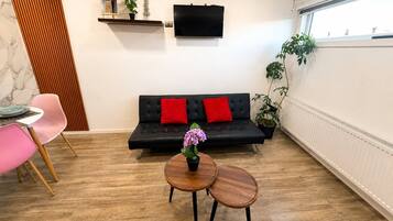 A modern living room with a black sofa, red cushions, a small wooden table with a vase, and a TV mounted on the wall.