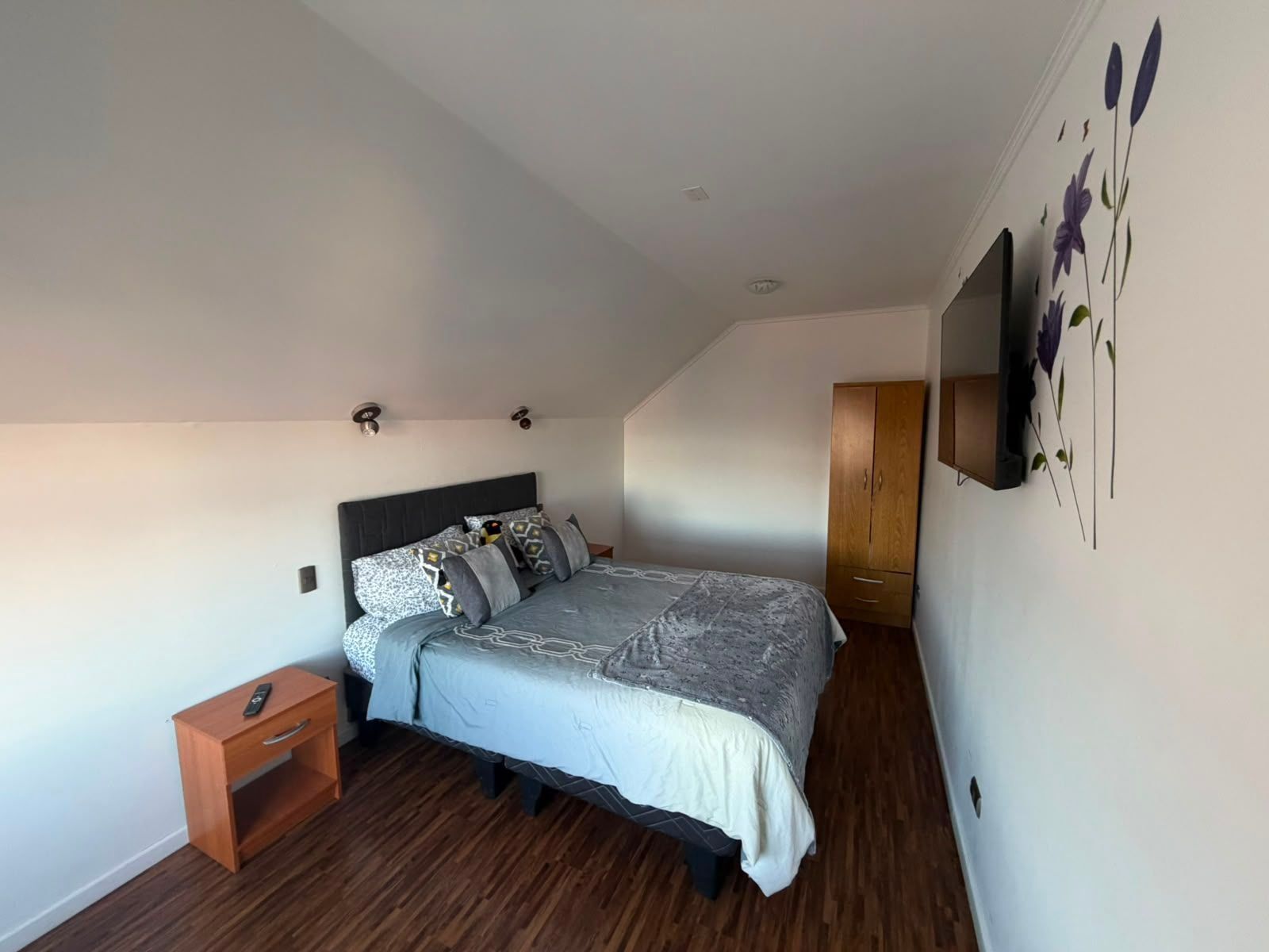 Un dormitorio con cama, mesita de noche, armario y televisor en la pared.