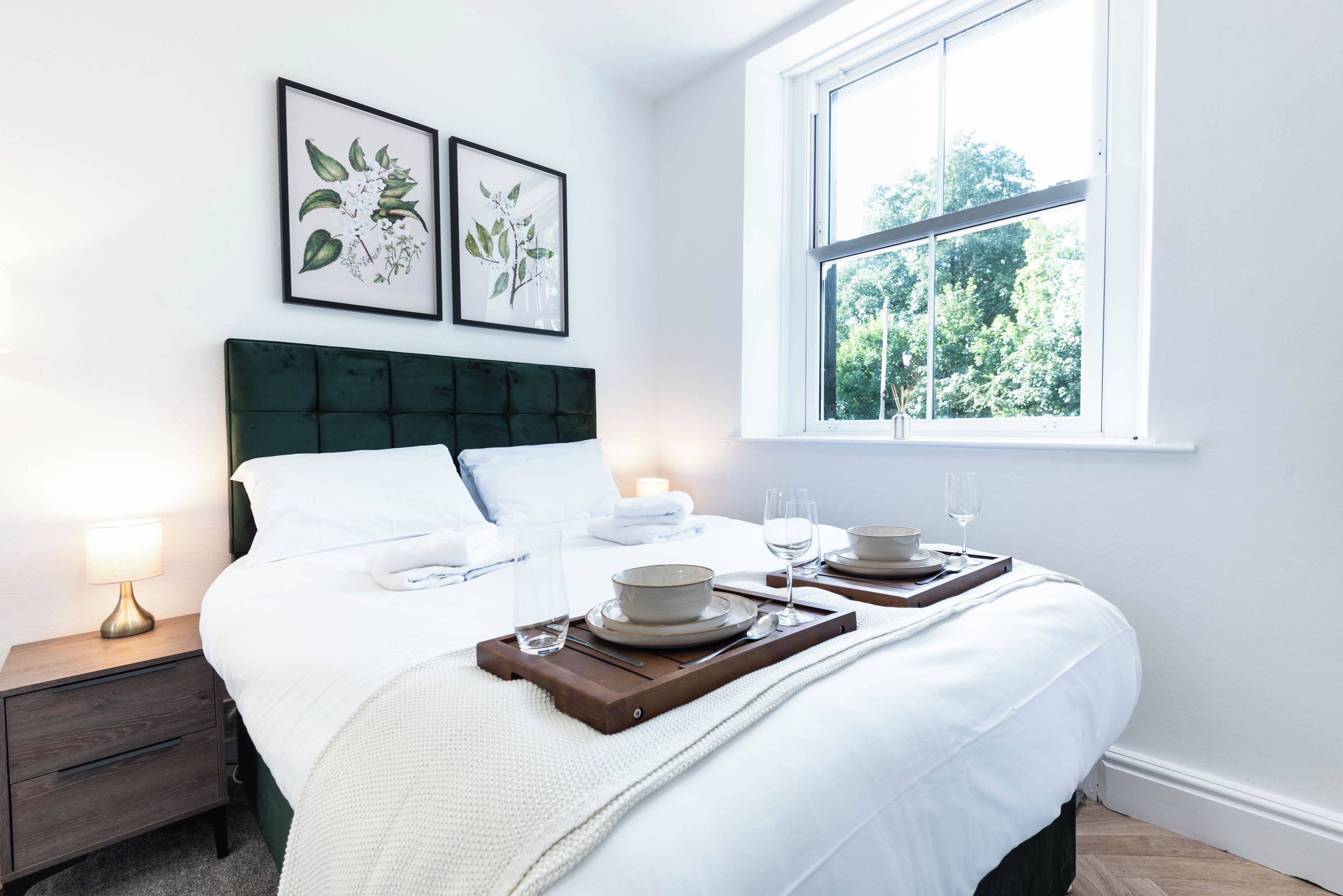 A neatly made bed with white linens, a dark green headboard, and a bedside table with a lamp. There are two framed botanical prints on the wall, and a window with a view of trees.