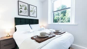 A neatly made bed with white linens, a dark green headboard, and a bedside table with a lamp. There are two framed botanical prints on the wall, and a window with a view of trees.