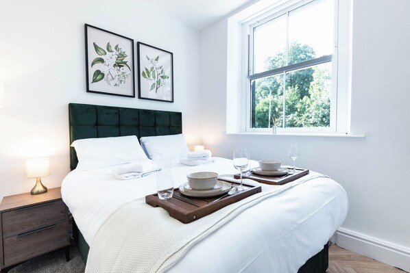 A neatly made bed with white linens, a dark green headboard, and a bedside table with a lamp. There are two framed botanical prints on the wall, and a window with a view of trees.