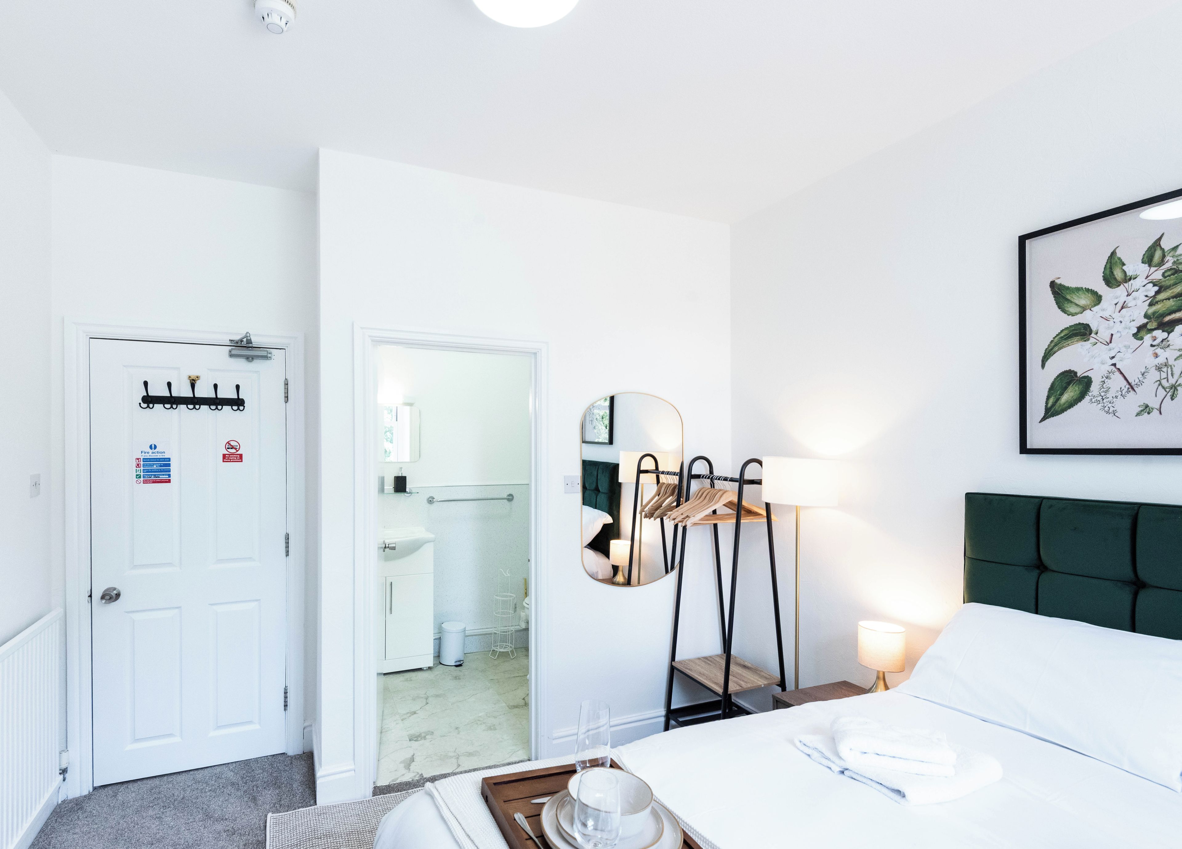 Ett hotellrum med en säng, ett sängbord med en lampa, en spegel, en klädställning och en dörr med en brandvarnare.
