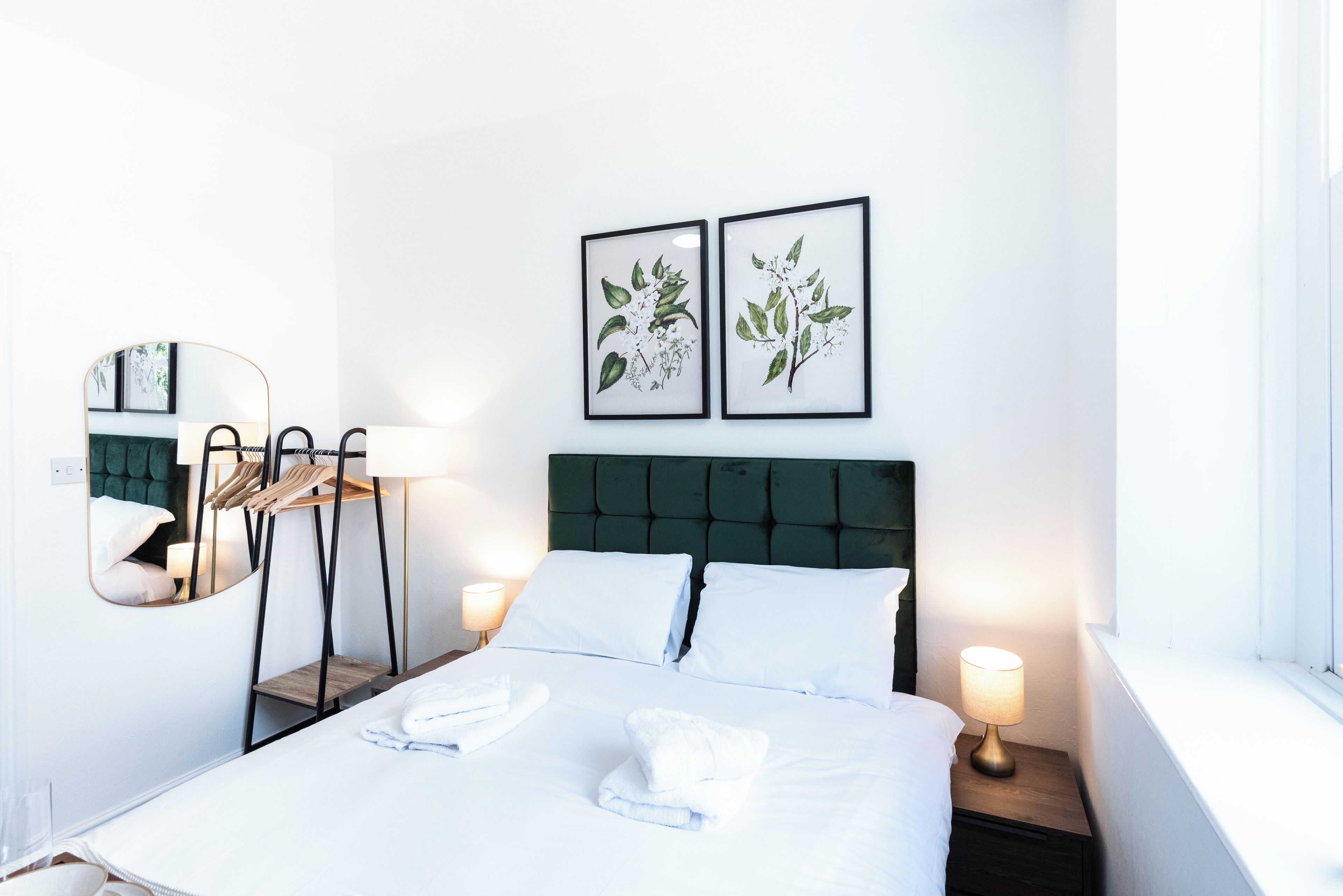 A hotel room with a bed, two bedside lamps, a mirror, a wooden side table, and two framed botanical prints on the wall.