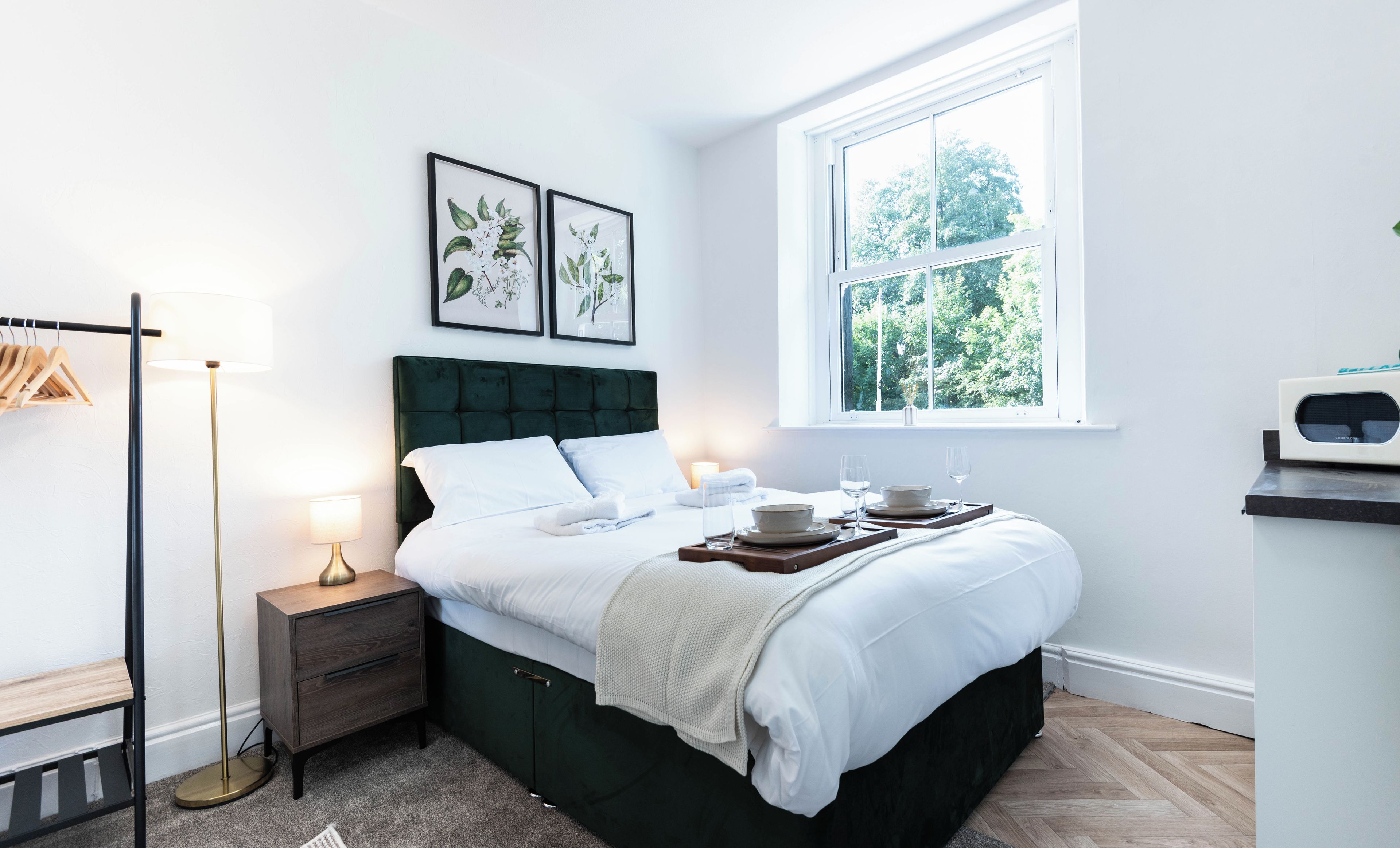 Une chambre à coucher bien rangée, avec un lit, des tables de chevet, une lampe sur pied et une fenêtre donnant sur des arbres.