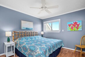 A bedroom with a bed, a nightstand, a chair, a ceiling fan, a window, and two paintings on the wall. - Casa De Ava: Ocean Views and Easy Beach Access (Oak Island)