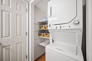A laundry room with a stacked washer and dryer, shelves with baskets, and a door. - Casa De Ava: Ocean Views and Easy Beach Access (Oak Island)
