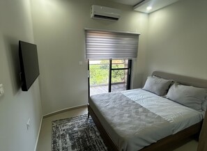 Un dormitorio con cama, televisión instalada en la pared, ventana con persianas y equipo de aire acondicionado.