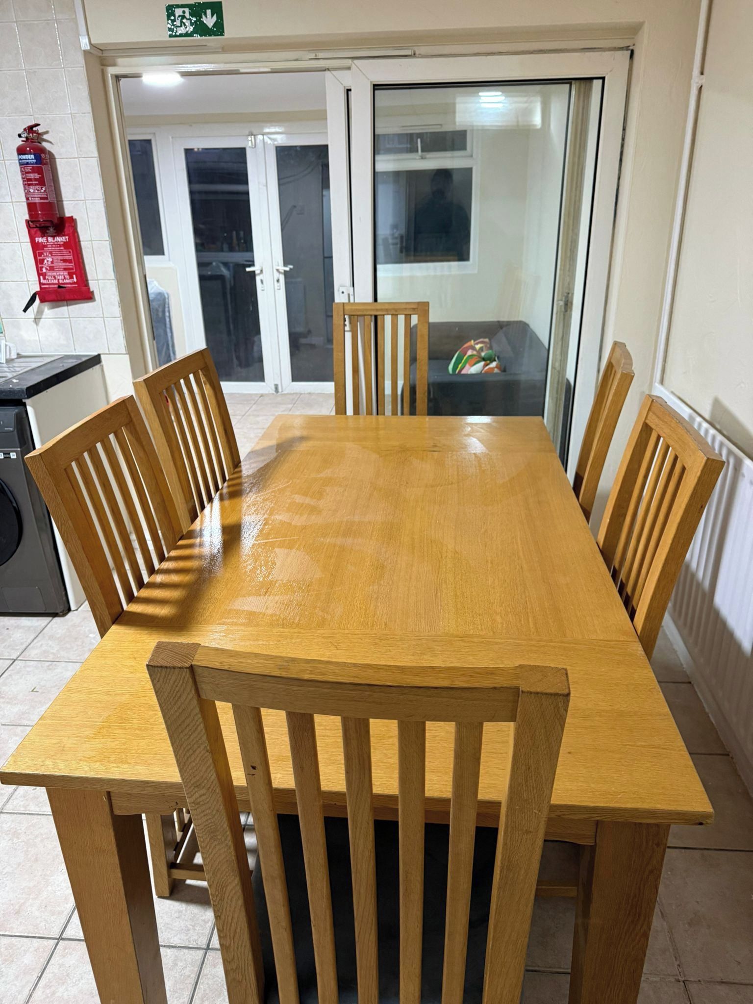 A wooden dining table with six chairs, a fire extinguisher, and a fire blanket on the wall.