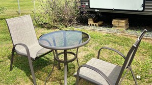 A recreational vehicle with a white panel, a glass table, and two chairs in a grassy area.